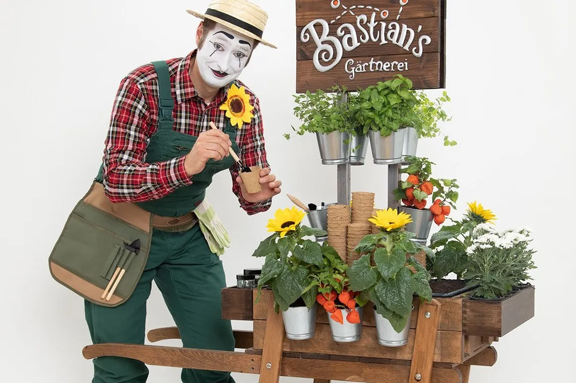 Ein Pantomime-Künstler mit weiß-schwarzem Gesichtsschminke und Strohhut beugt sich über einen Holzwagen mit Pflanzen, Töpfen und Gärtnerwerkzeug. Im Hintergrund ein Schild mit der Aufschrift 