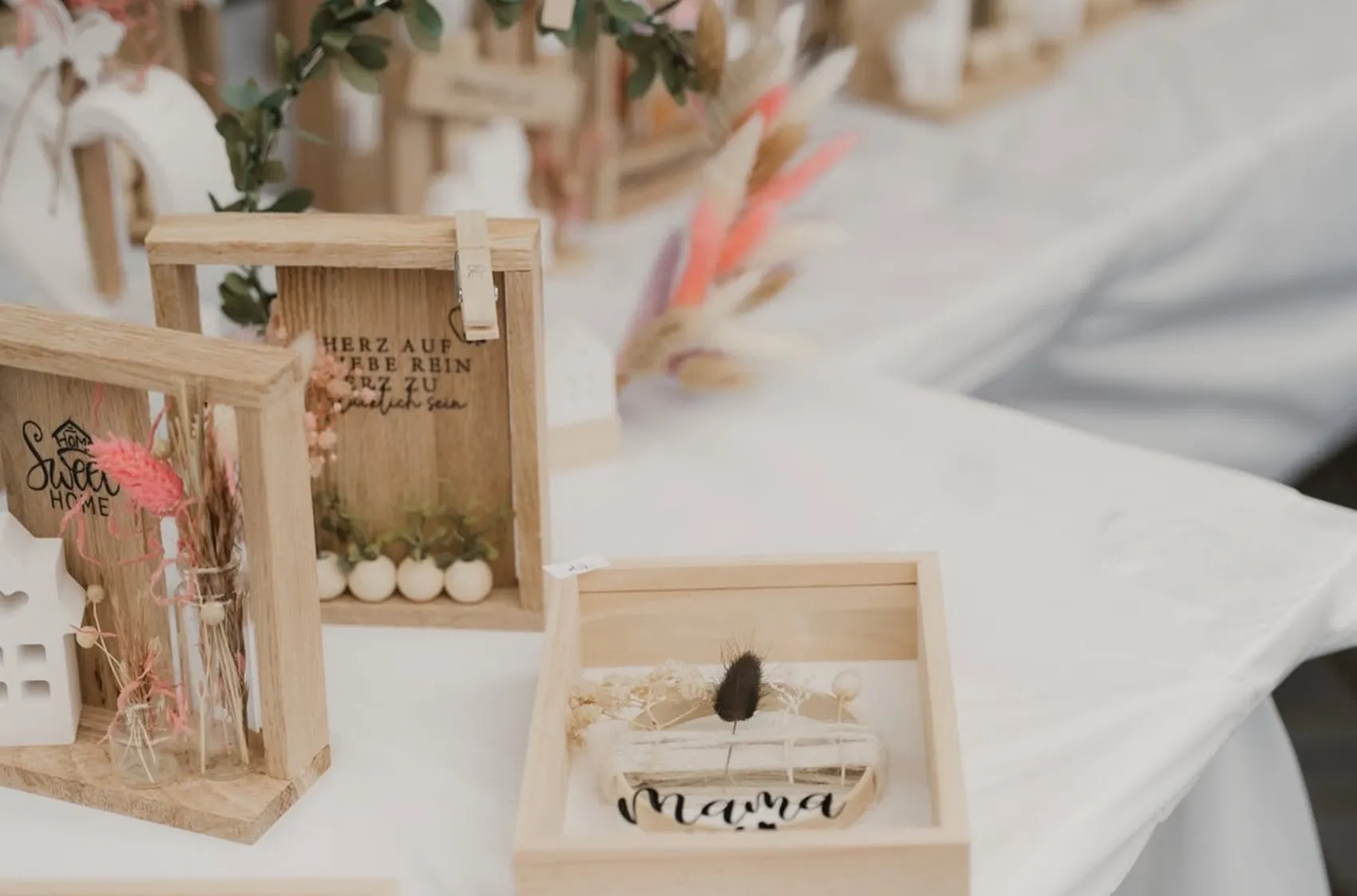 Holzrahmen mit Dekorationen und Schriftzügen wie „Sweet Home“ und „Mama“ stehen auf einem weißen Tisch.