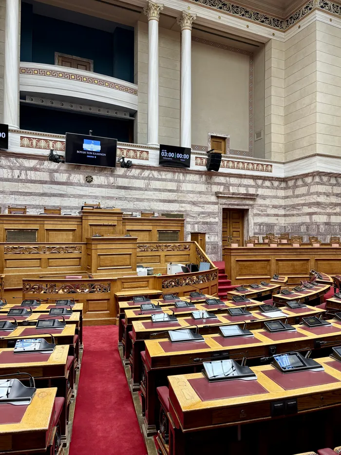 Innenansicht des griechischen Parlaments in Athen mit hölzernen Sitzreihen, Marmorwänden und großen weißen Säulen.