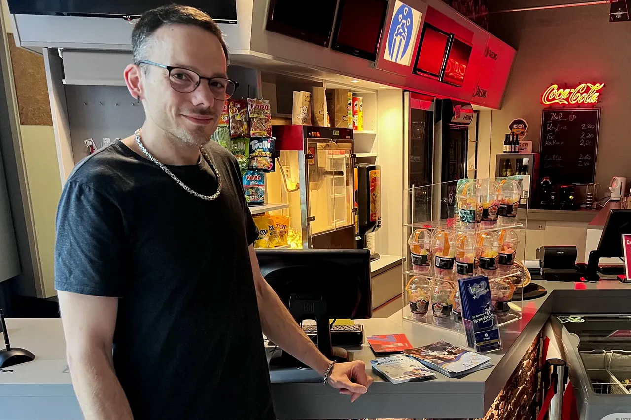 Mann mit Brille und Kappe steht lächelnd hinter einer Theke in einem Kinosaal, umgeben von Getränken und Snacks.