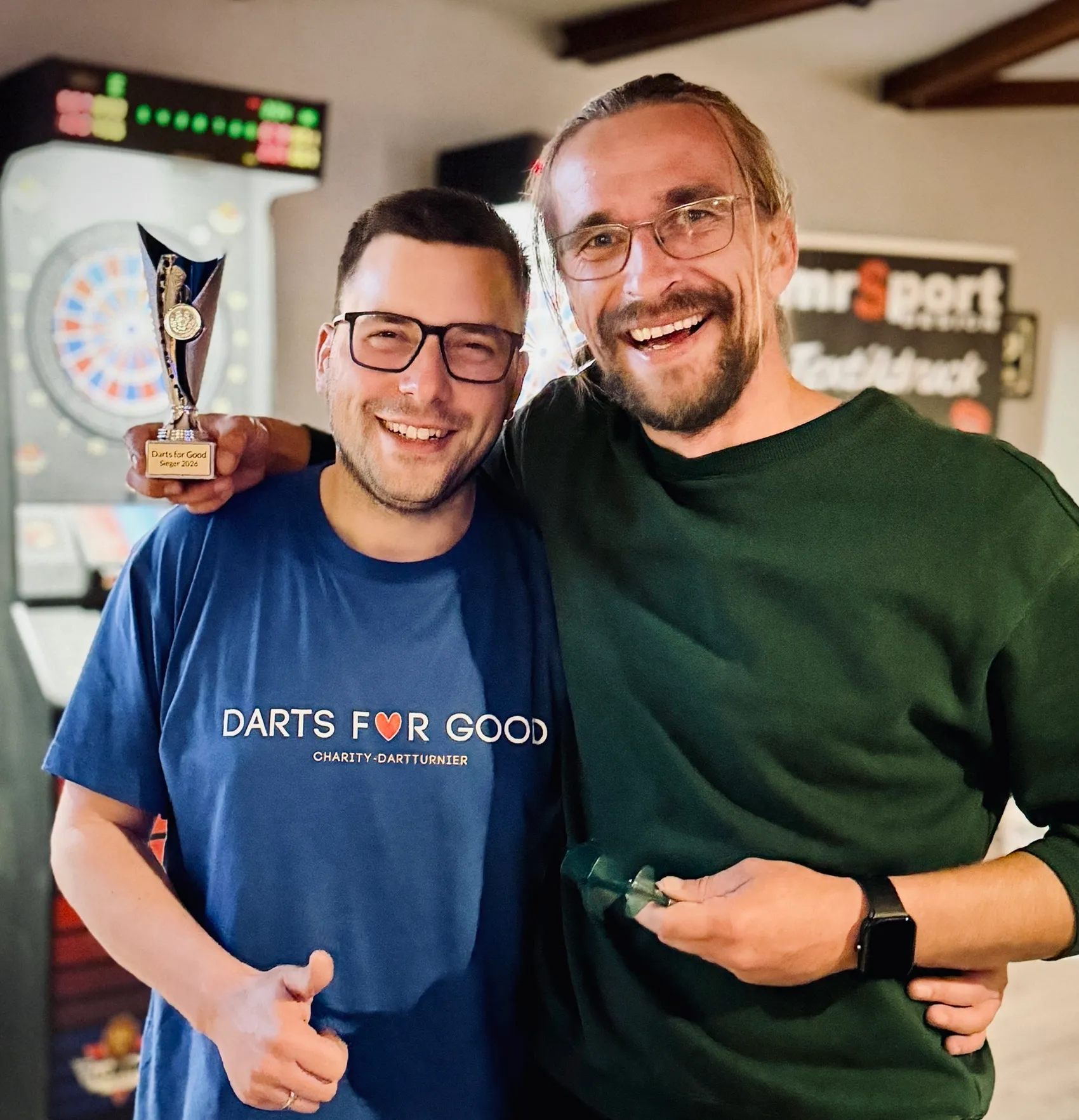 Patrick Jahn und Lothar Pniok posieren gemeinsam mit der Trophäe des Benefiz-Dartturniers „Darts for Good“.