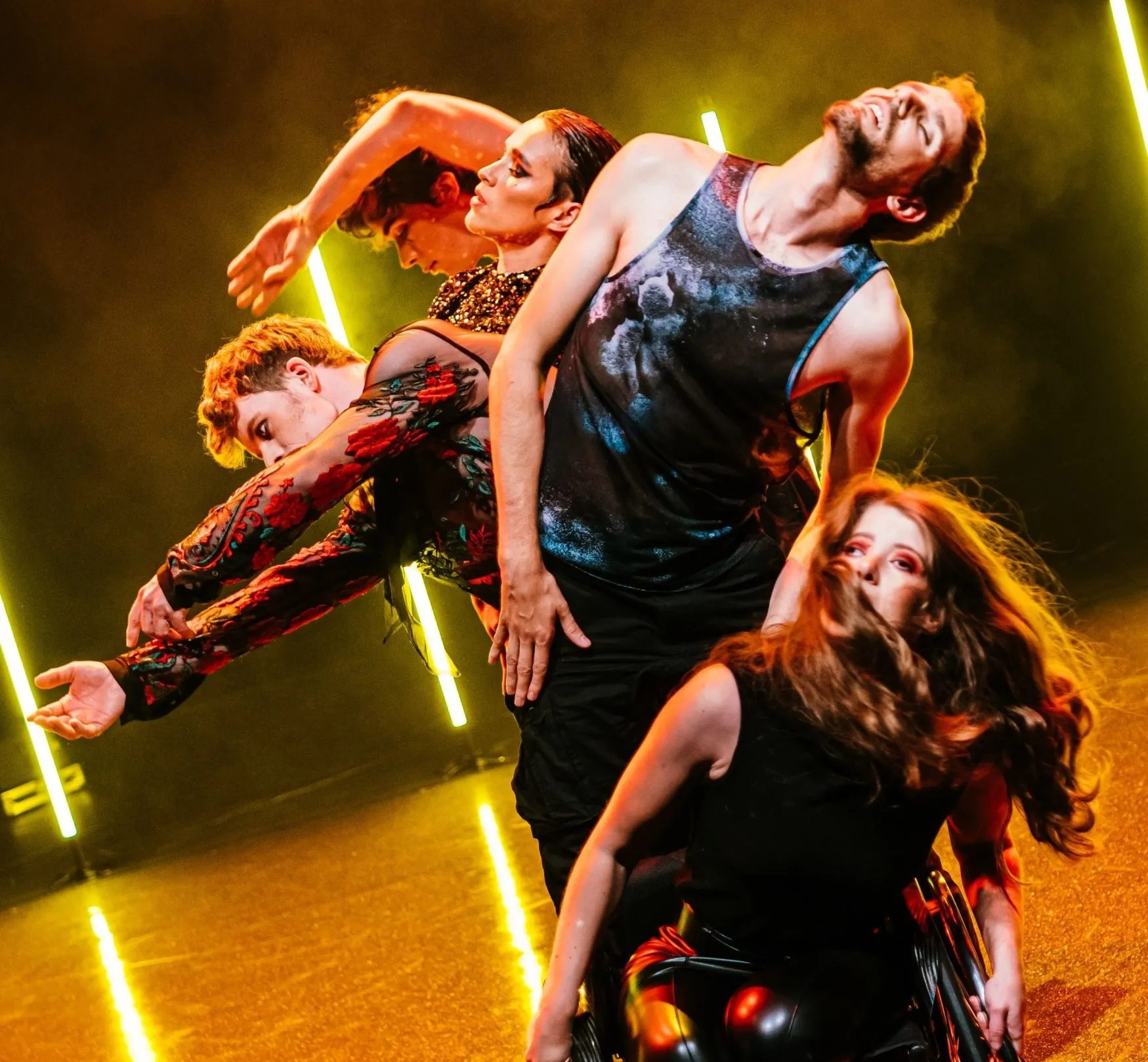 Vier Tänzer*innen in Bewegung, eine davon im Rollstuhl, vor gelbem Licht und Neonröhren. Die Performance betont künstlerische Teilhabe und Vielfalt.