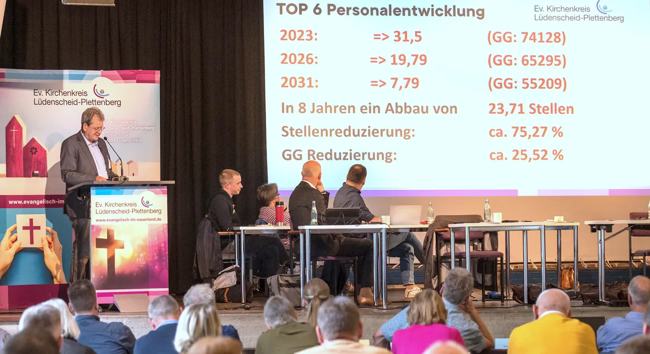 Martin Pogorzelski präsentiert vor einem Publikum von Synodalen eine Grafik zur Personalentwicklung des Kirchenkreises Lüdenscheid-Plettenberg, die einen deutlichen Stellenabbau bis 2031 zeigt. Im Hintergrund sind Kreuze und das Logo des Kirchenkreises zu sehen.
