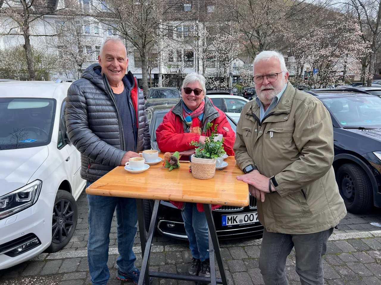 Drei ältere Menschen stehen auf einem Parkplatz vor mehreren Autos und unterhalten sich. Ein Mann hält eine Pflanze in einem Korb.