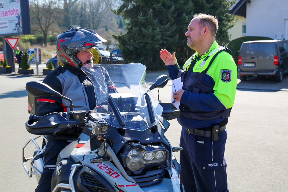 Polizist erklärt Motorradfahrer etwas. Im Hintergrund ein Parkplatz und ein Auto.