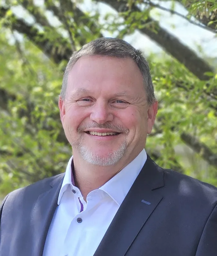 Porträt von Peter Schrade, einem Mann mittleren Alters mit grau meliertem Haar und Bart, der in einen Anzug gekleidet ist und lächelt.
