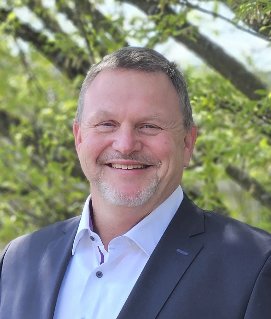 Porträt von Peter Schrade, einem Mann mittleren Alters mit grau meliertem Haar und Bart, der in einen Anzug gekleidet ist und lächelt.