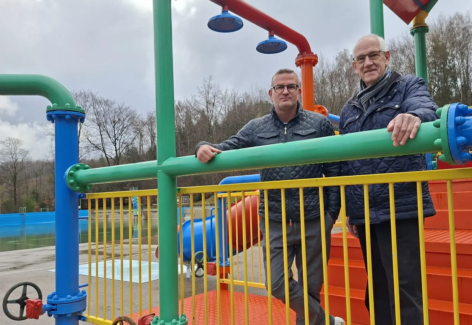 Zwei Männer stehen vor einem bunten Wasserspielplatz mit Rohren und Ventilen. Im Hintergrund ein Becken und Bäume.