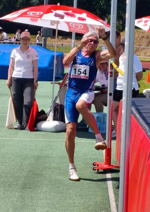 Ein älterer Mann in Sportkleidung springt beim Hochsprung über eine Latte. Im Hintergrund stehen weitere Sportler und eine Schiedsrichterin.