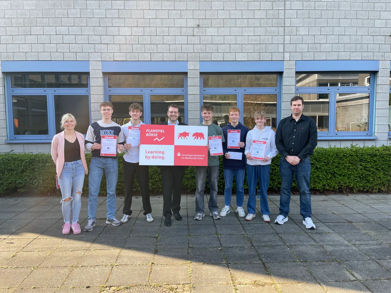 Sieben Schüler und ein Mann stehen lächelnd vor einem Schulgebäude und halten Urkunden. Im Hintergrund ein Banner mit dem Aufdruck „Planspiel Börse“ und dem Logo der Sparkasse.