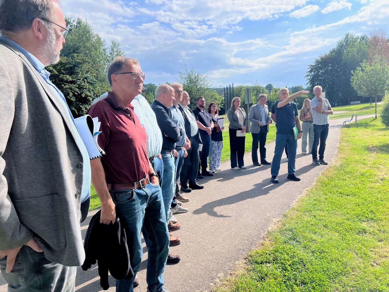 Eine Gruppe von Personen steht auf einem Gehweg neben einer Wiese und diskutiert, während eine Person in Richtung eines Feldes deutet.