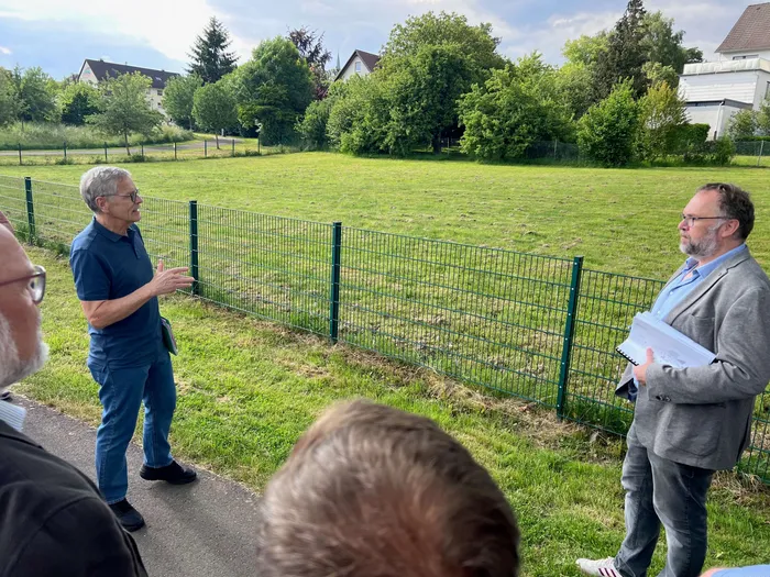 Zwei Männer erklären vor einer Grünfläche und einem Maschendrahtzaun Pläne, während eine kleine Gruppe zuhört. Im Hintergrund sind Wohnhäuser zu sehen.