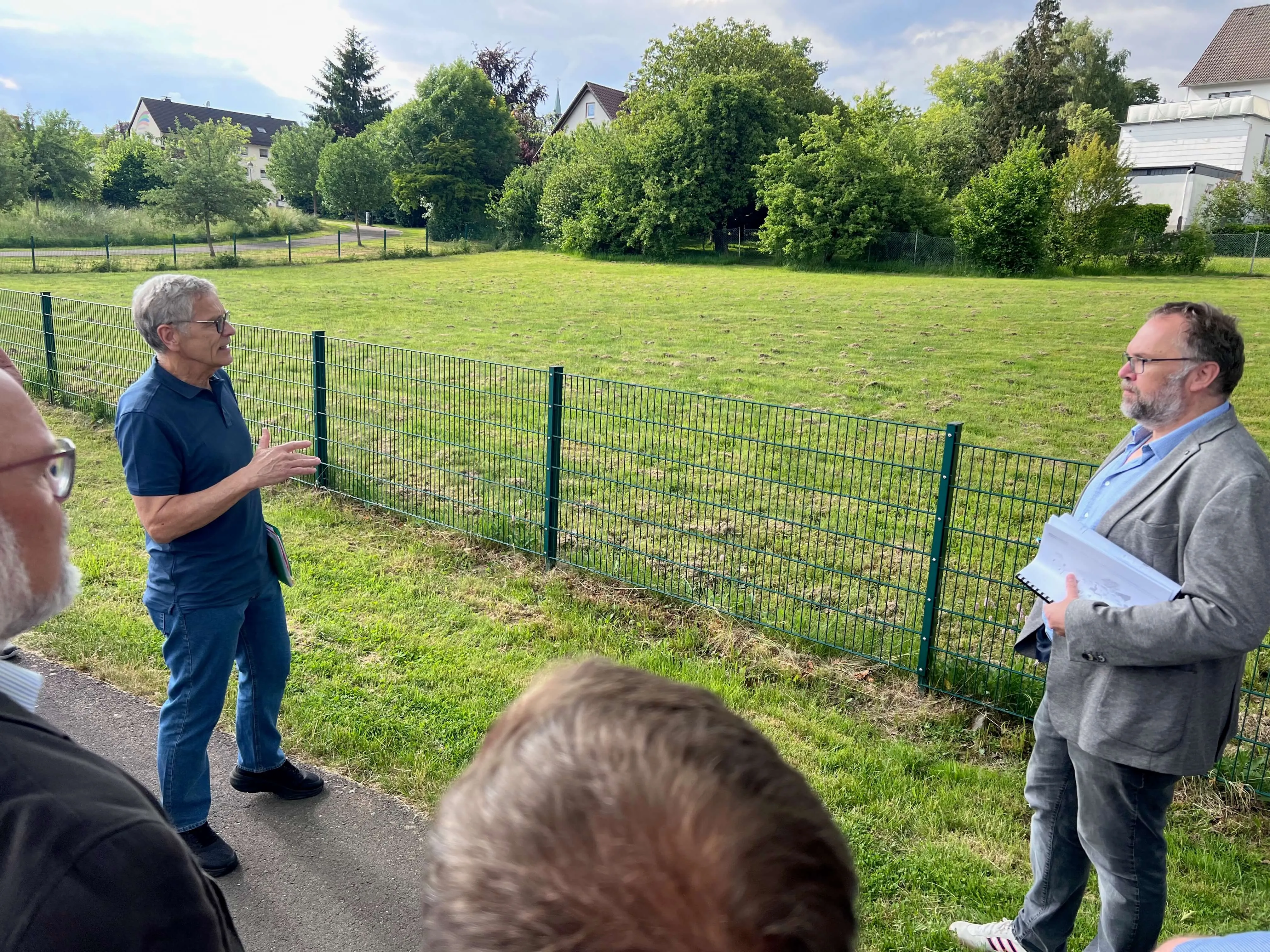 Zwei Männer erklären vor einer Grünfläche und einem Maschendrahtzaun Pläne, während eine kleine Gruppe zuhört. Im Hintergrund sind Wohnhäuser zu sehen.