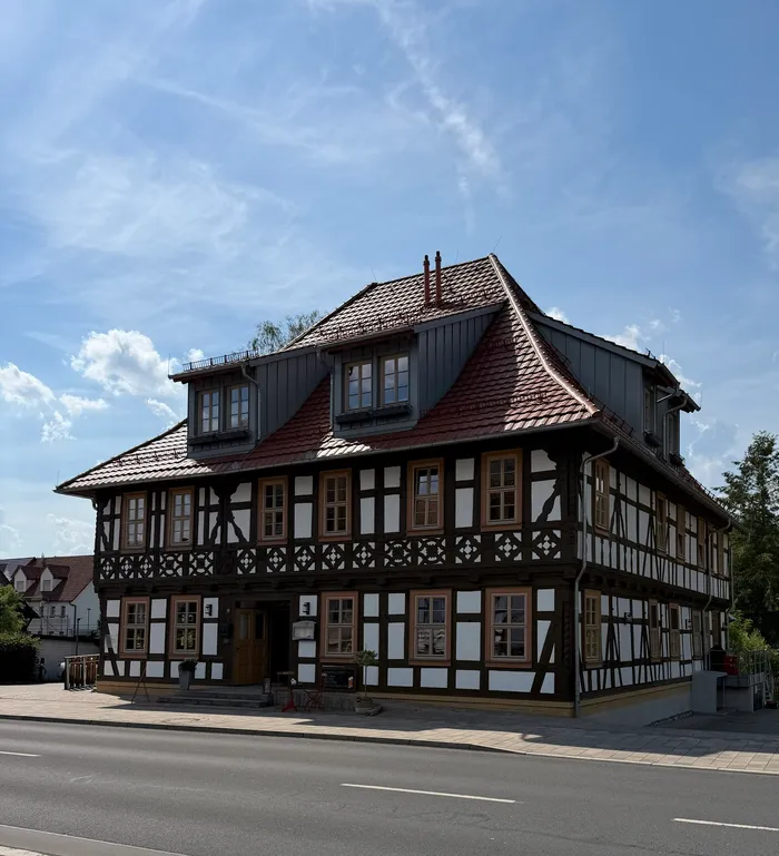 Ein zweistöckiges Fachwerkhaus mit rotem Ziegeldach steht an einer Straße. Im Erdgeschoss sind mehrere Fenster und eine Tür zu sehen.