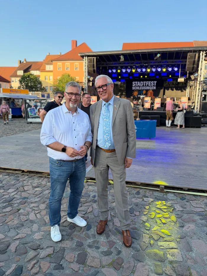 Zwei Männer im Anzug und legerer Kleidung stehen lächelnd vor einer Bühne mit der Aufschrift „Stadtfest Schleusingen“. Im Hintergrund sind weitere Personen und historische Gebäude zu sehen.