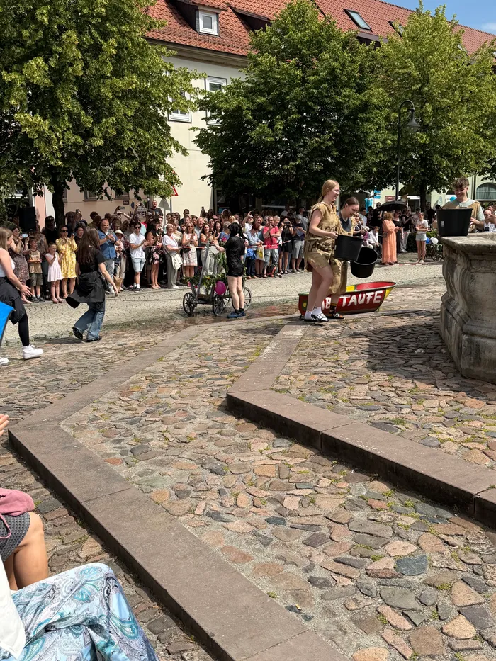 Eine Frau spricht in ein Mikrofon auf einer erhöhten Fläche, während im Hintergrund eine Menschenmenge auf einer Pflasterstraße steht. Im Hintergrund sind Gebäude und Bäume zu sehen.