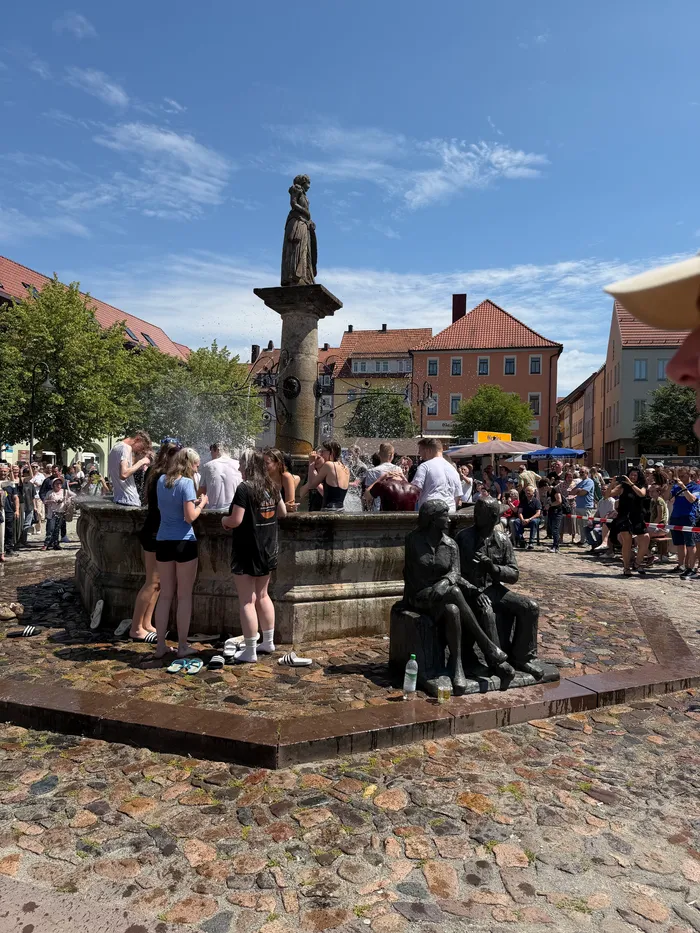 Menschen stehen und sitzen um einen historischen Brunnen auf einem Platz mit Kopfsteinpflaster. Im Hintergrund sind Gebäude und ein blauer Himmel mit wenigen Wolken zu sehen.