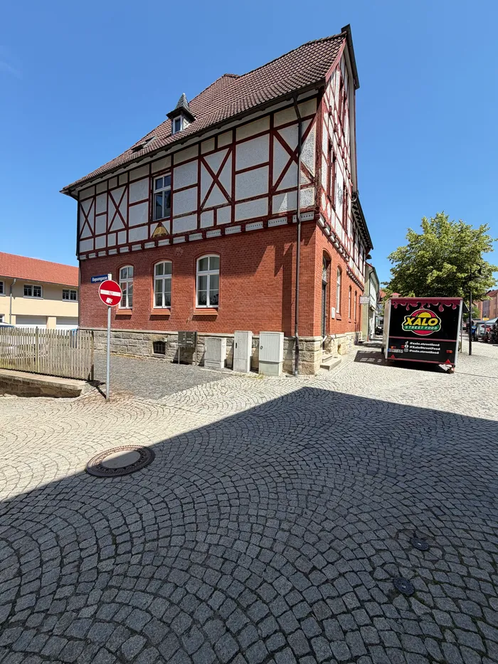 Ein zweistöckiges Fachwerkhaus mit roten Backsteinen und einem dunklen Satteldach steht an einer Straßenecke. Vor dem Haus befindet sich ein Schild für 