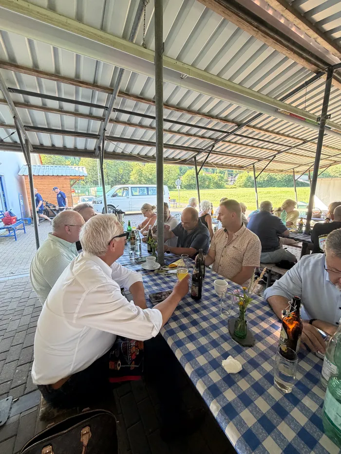 Eine Gruppe von Menschen sitzt an einem Tisch unter einem Sonnensegel, Getränke und Snacks sind darauf verteilt. Im Hintergrund sind weitere Personen und ein Gebäude zu sehen.