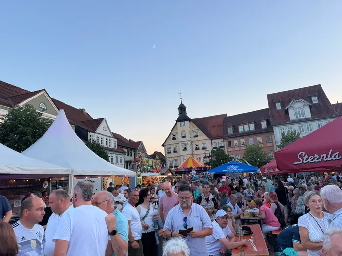 Eine Menschenmenge steht auf einem Stadtplatz vor Fachwerkhäusern und feiert. Im Vordergrund sind Personen unterschiedlichen Alters zu sehen, einige halten Getränke in der Hand. Im Hintergrund ein Kirchturm und ein blauer Himmel mit einem Halbmond.