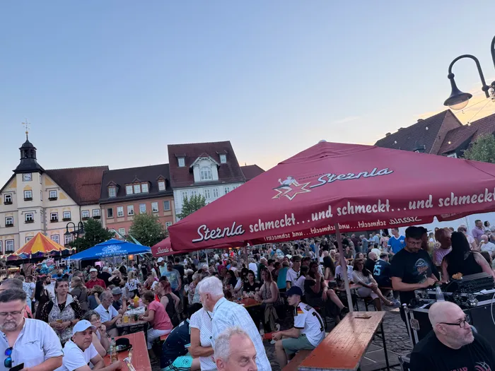 Eine Menschenmenge sitzt vor einem Gebäude mit einem großen Werbebanner für Sternla-Bier. Im Hintergrund sind weitere Gebäude und ein Kirchturm zu sehen.
