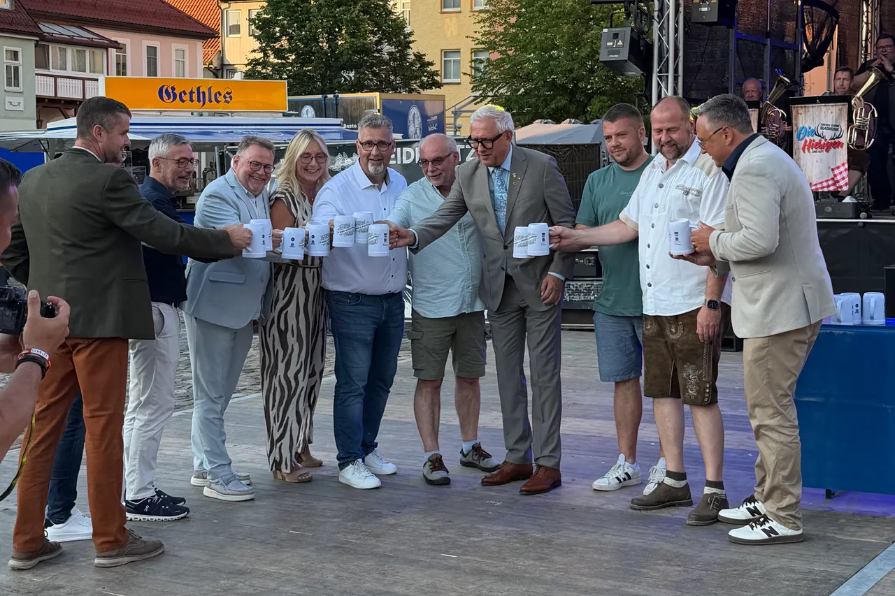 Eine Gruppe von neun Personen, darunter Bürgermeister und Stadträte, hält Bierkrüge in der Hand und lächelt in die Kamera. Im Hintergrund ist eine Bühne mit der Aufschrift „Die Heidelberger“ zu sehen.