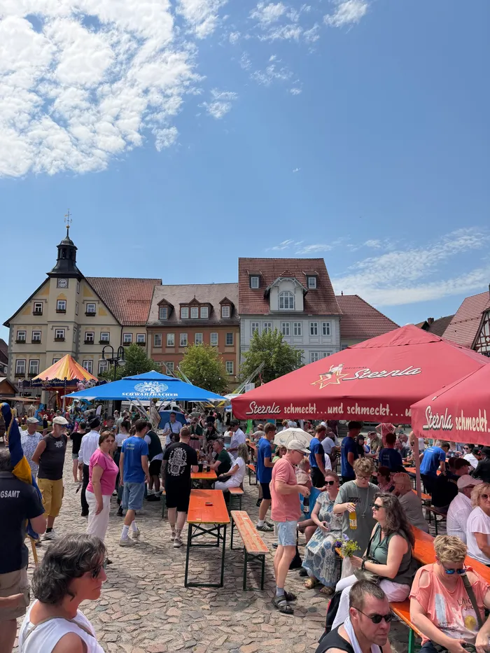 Menschen stehen auf einem Platz vor Fachwerkhäusern und unter roten Sonnenschirmen mit der Aufschrift „Sternla“. Im Hintergrund ein Kirchturm und ein blauer Himmel mit wenigen Wolken.