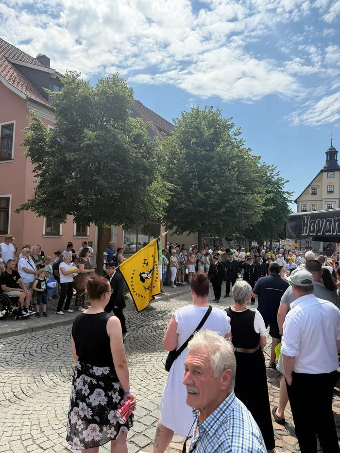 Eine Menschenmenge steht auf einem Platz vor Fachwerkhäusern. Im Vordergrund hält eine Person eine gelbe Fahne mit der Aufschrift „Partnerschaft Plettenberg-Schleusingen“.