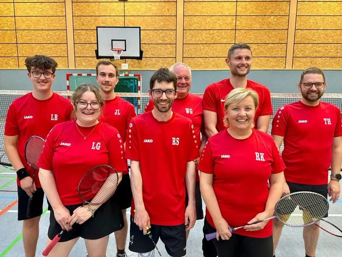 Sieben Mitglieder der ersten PBV-Badmintonmannschaft in roten Sportshirts posieren mit Badmintonschlägern in einer Sporthalle.