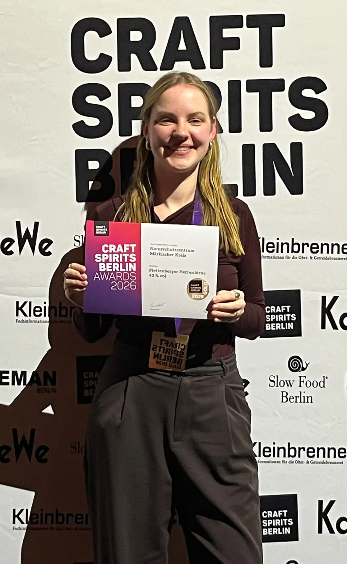 Johanna Knipp hält Goldmedaille und Urkunde des Craft Spirits Berlin Awards 2026 für Plettenberger Herrenbirne vor einem Hintergrund mit dem Schriftzug „Craft Spirits Berlin“.