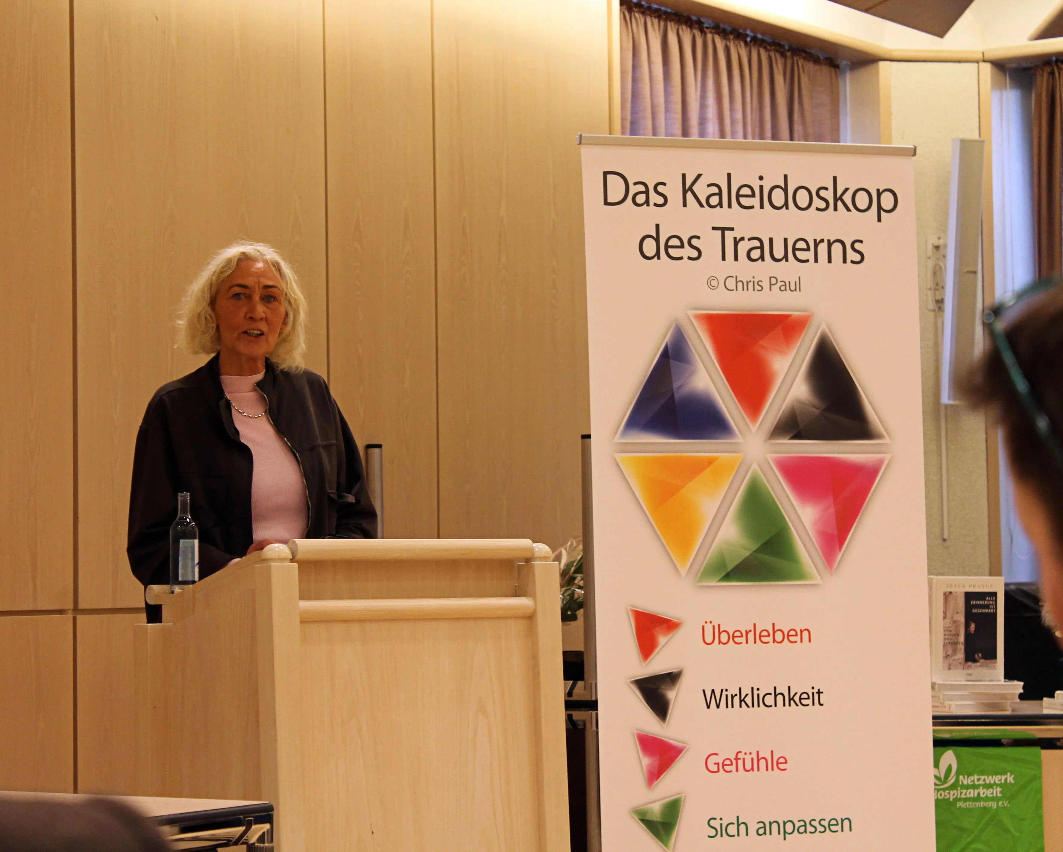 Frau mit grauen Haaren spricht vor einem Roll-up-Banner mit dem Titel „Das Kaleidoskop des Trauerns“ und farbigen Dreiecken, die die Begriffe „Überleben“, „Wirklichkeit“, „Gefühle“ und „Sich anpassen“ darstellen.