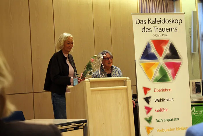 Zwei Frauen an einem Rednerpult, im Hintergrund ein Roll-up-Banner mit dem Titel „Das Kaleidoskop des Trauerns“ und farbigen Segmenten, die Themen wie „Überleben“, „Wirklichkeit“ und „Gefühle“ darstellen.