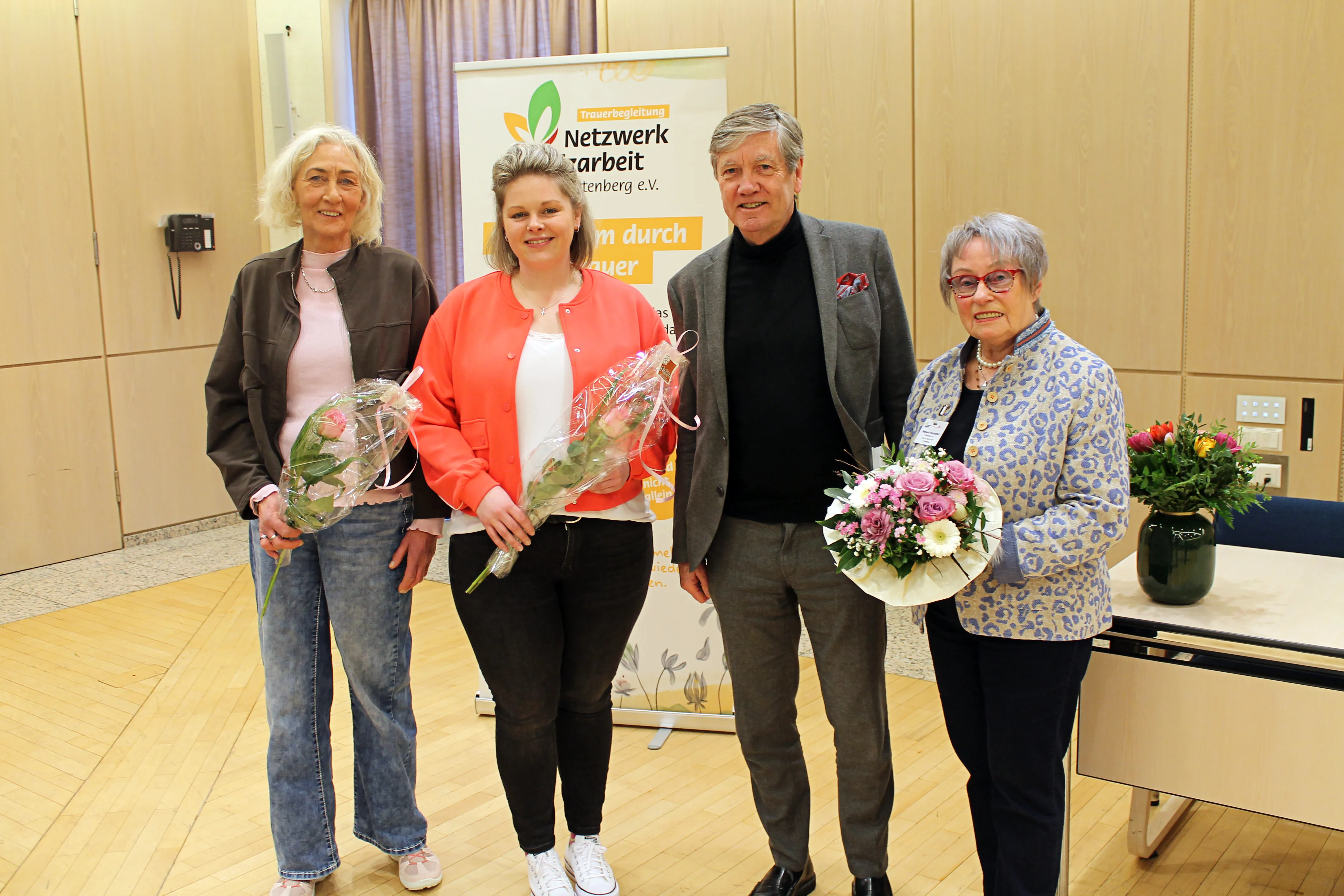 Vier Personen halten Blumensträuße vor einem Banner mit dem Aufdruck „Trauerbegleitung Netzwerk Sterben und Leben“.
