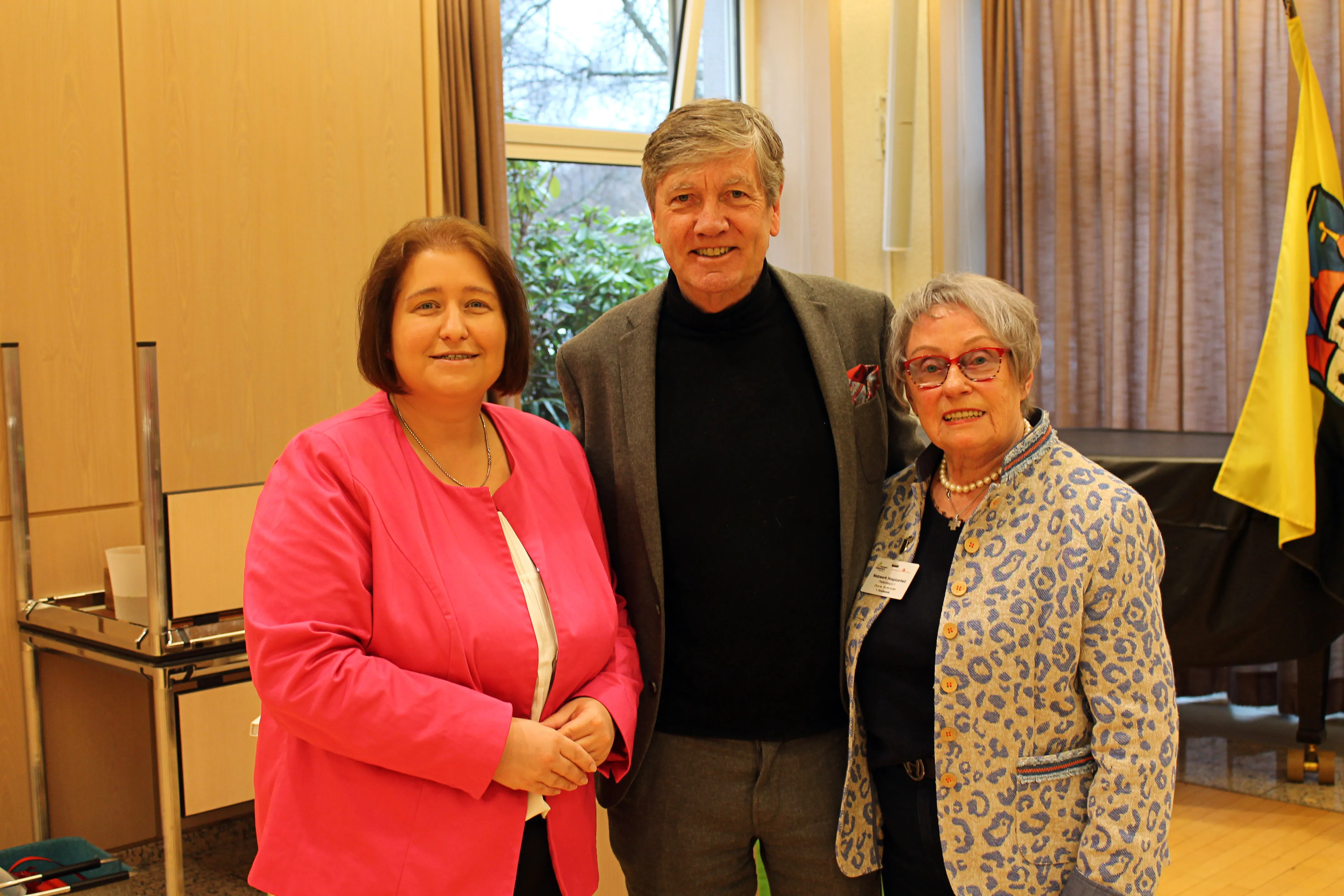 Drei Personen lächeln in die Kamera: eine Frau in einem pinken Blazer, ein Mann in einem grauen Anzug und eine ältere Frau mit rotem Haar und einer gemusterten Jacke. Im Hintergrund sind Vorhänge und eine Flagge zu sehen.