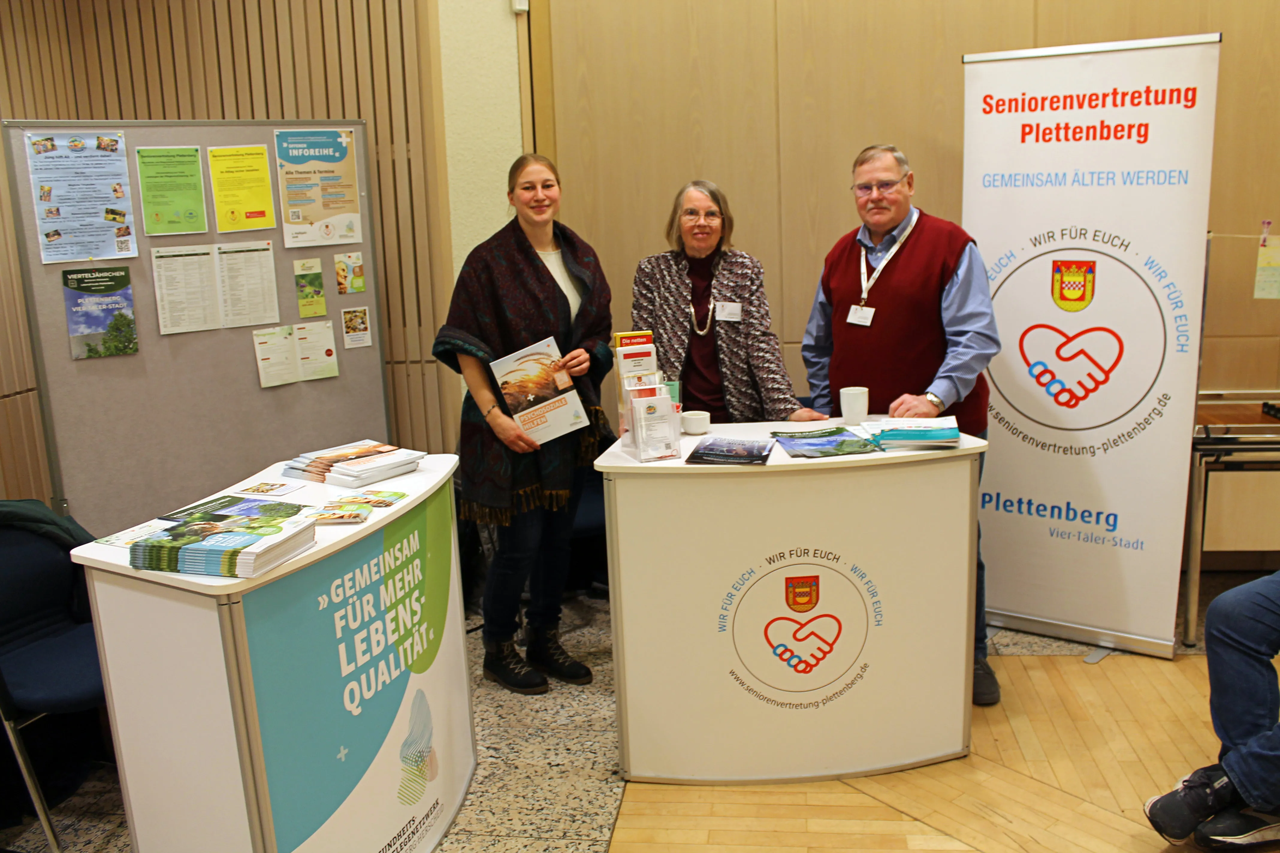 Drei ehrenamtliche Mitarbeiterinnen und Mitarbeiter des Seniorenvertretung Plettenberg stehen an einem Informationsstand mit Broschüren und Roll-ups. Im Hintergrund hängen Plakate und Flyer.