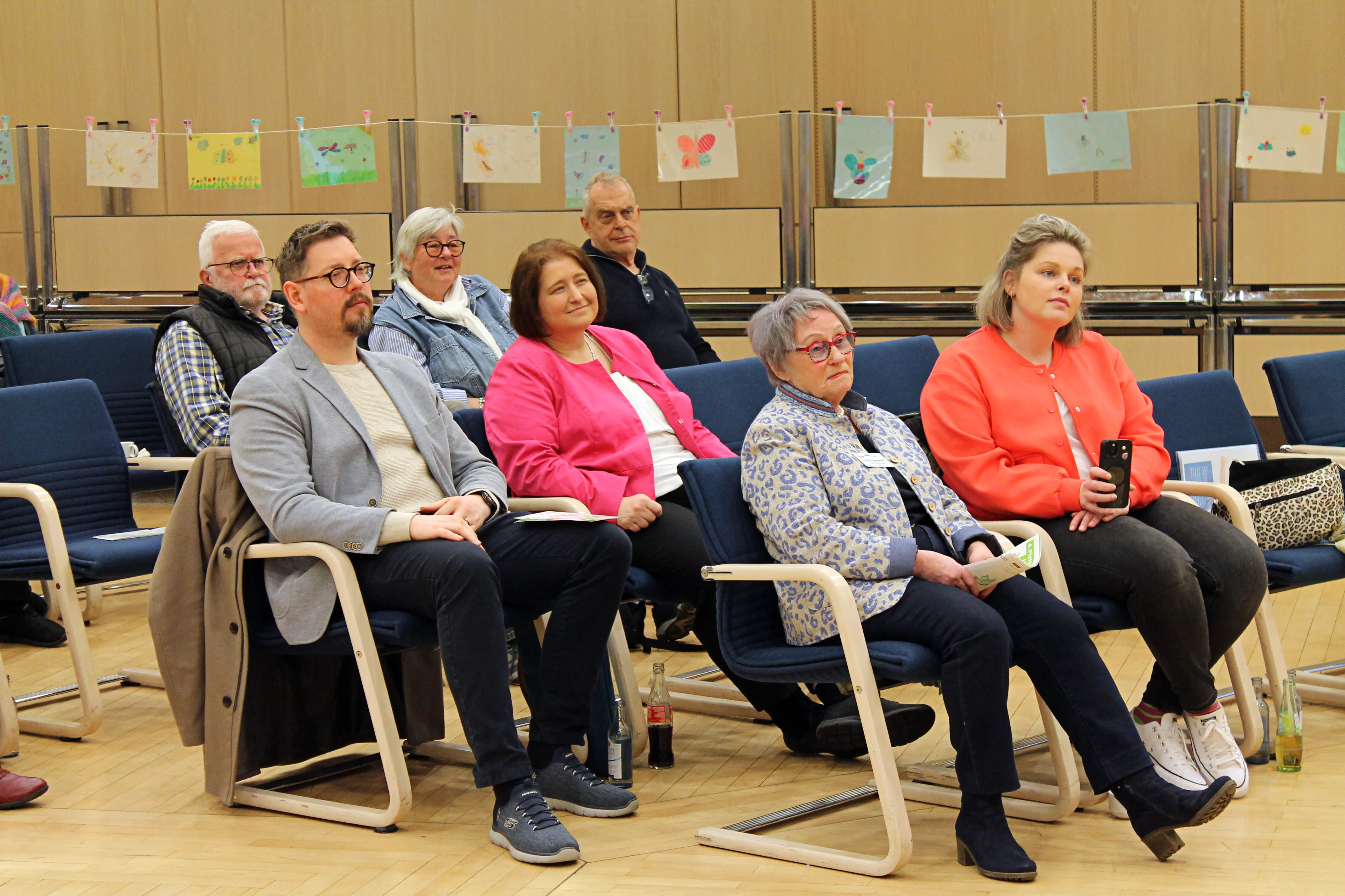 Zuschauer sitzen auf Stühlen in einem hellen Raum vor einer Wand mit bunten Kinderzeichnungen. Sie verfolgen aufmerksam eine Veranstaltung zum Thema Abschied und Erinnerung.