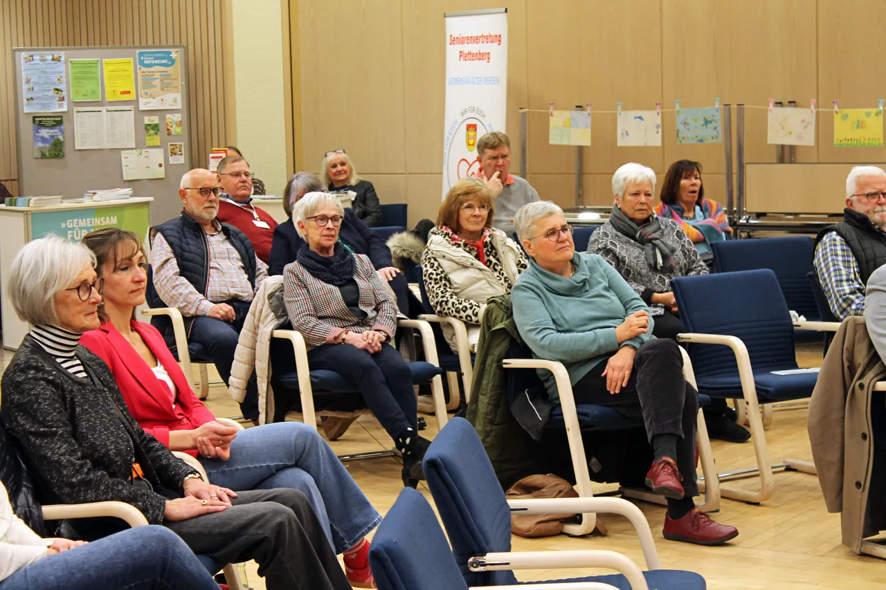 Zuschauer sitzen auf weißen Stühlen in einem hellen Raum und hören zu. Im Hintergrund ein Plakat mit dem Hinweis auf den Plettenberger Hospiztag.
