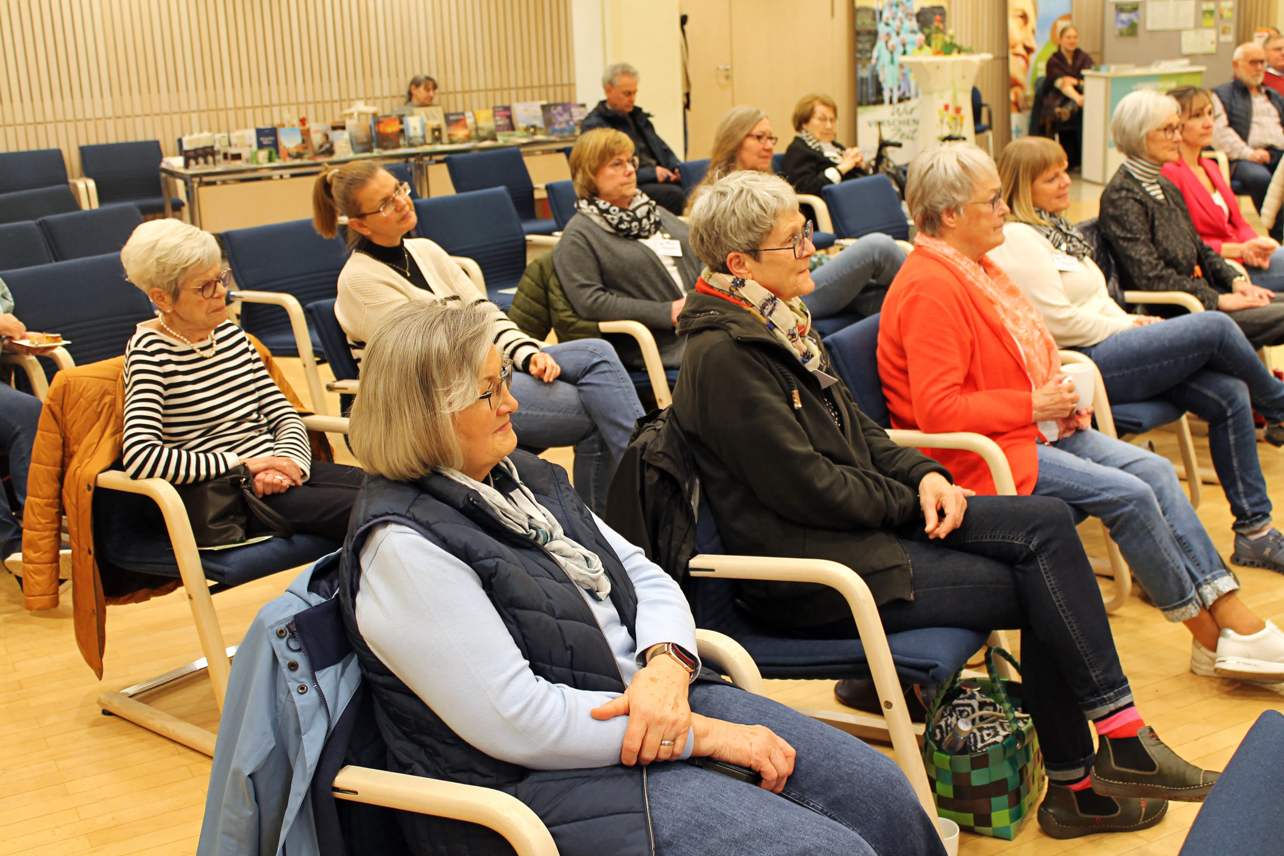 Zuschauer sitzen auf hellen Stühlen in einem Raum mit Holzvertäfelung und verfolgen eine Veranstaltung zum Thema Hospiz und Trauerbegleitung.