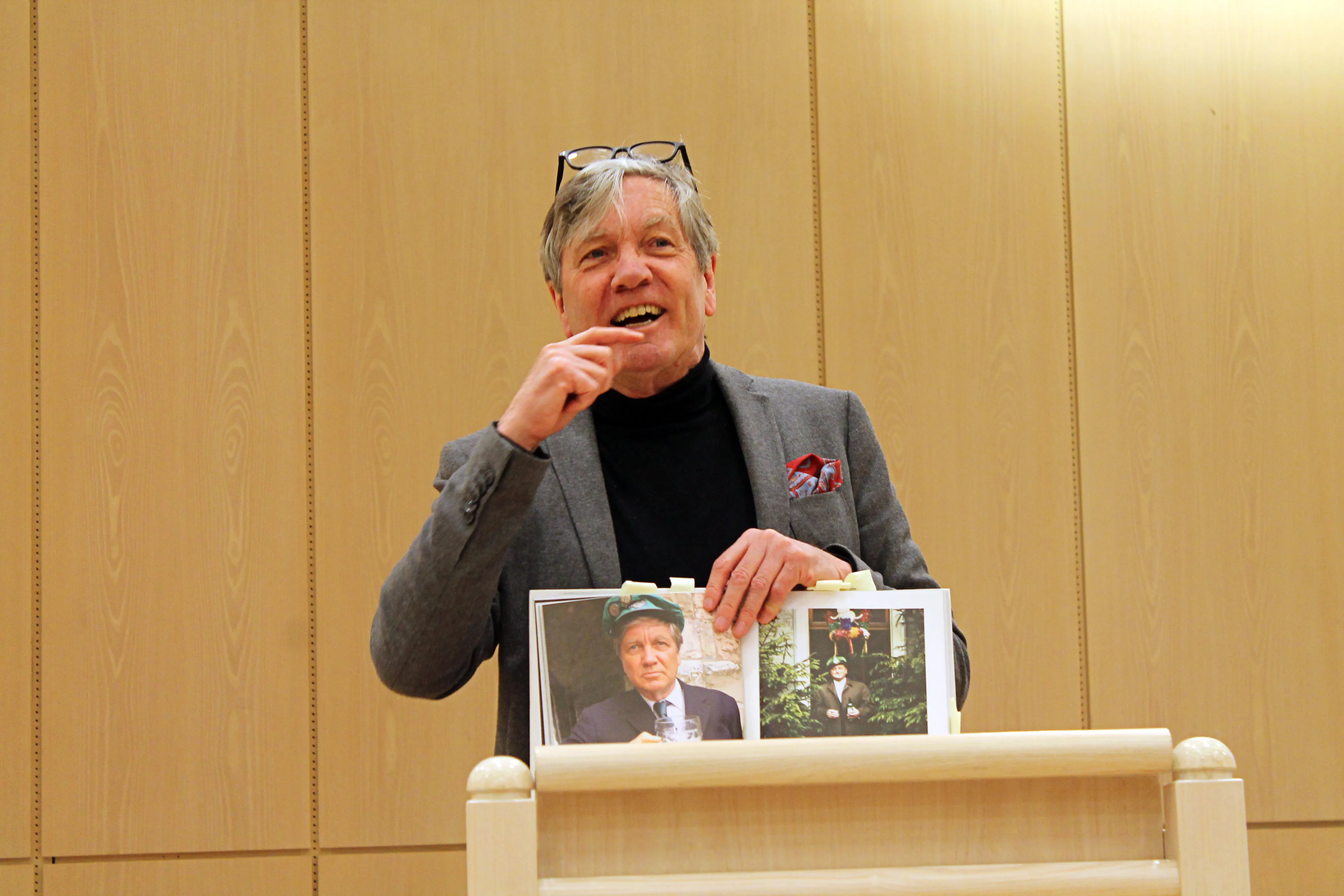 Ein älterer Mann mit Brille hält Fotos in der Hand und spricht fröhlich in die Menge. Im Hintergrund eine Holzvertäfelung.