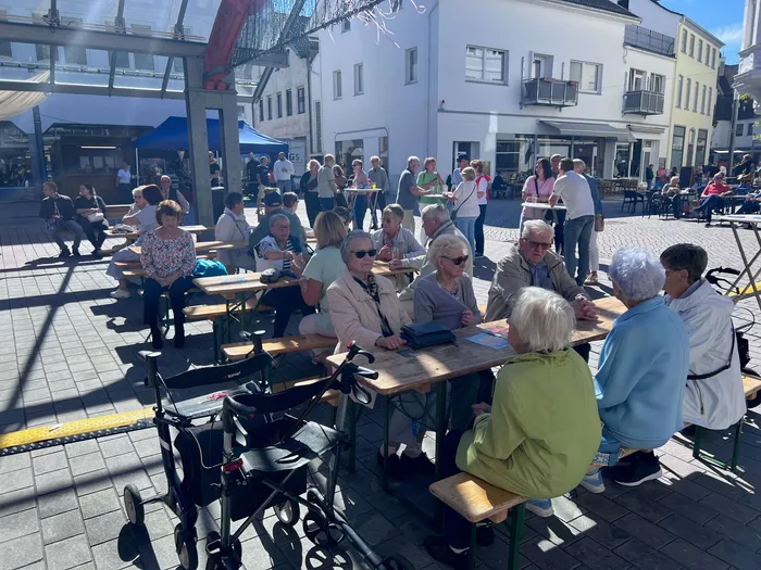 Eine Gruppe älterer Menschen sitzt an Holztischen auf dem sonnigen Alten Markt; im Vordergrund steht ein Rollator.