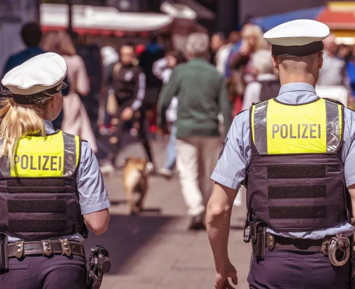Zwei Polizisten mit gelben Warnwesten und der Aufschrift „POLIZEI“ gehen mit dem Rücken zum Betrachter durch eine Menschenmenge.