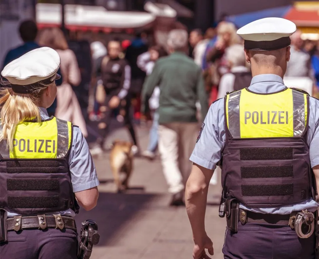 Zwei Polizisten mit gelben Warnwesten und der Aufschrift „POLIZEI“ gehen mit dem Rücken zum Betrachter durch eine Menschenmenge.