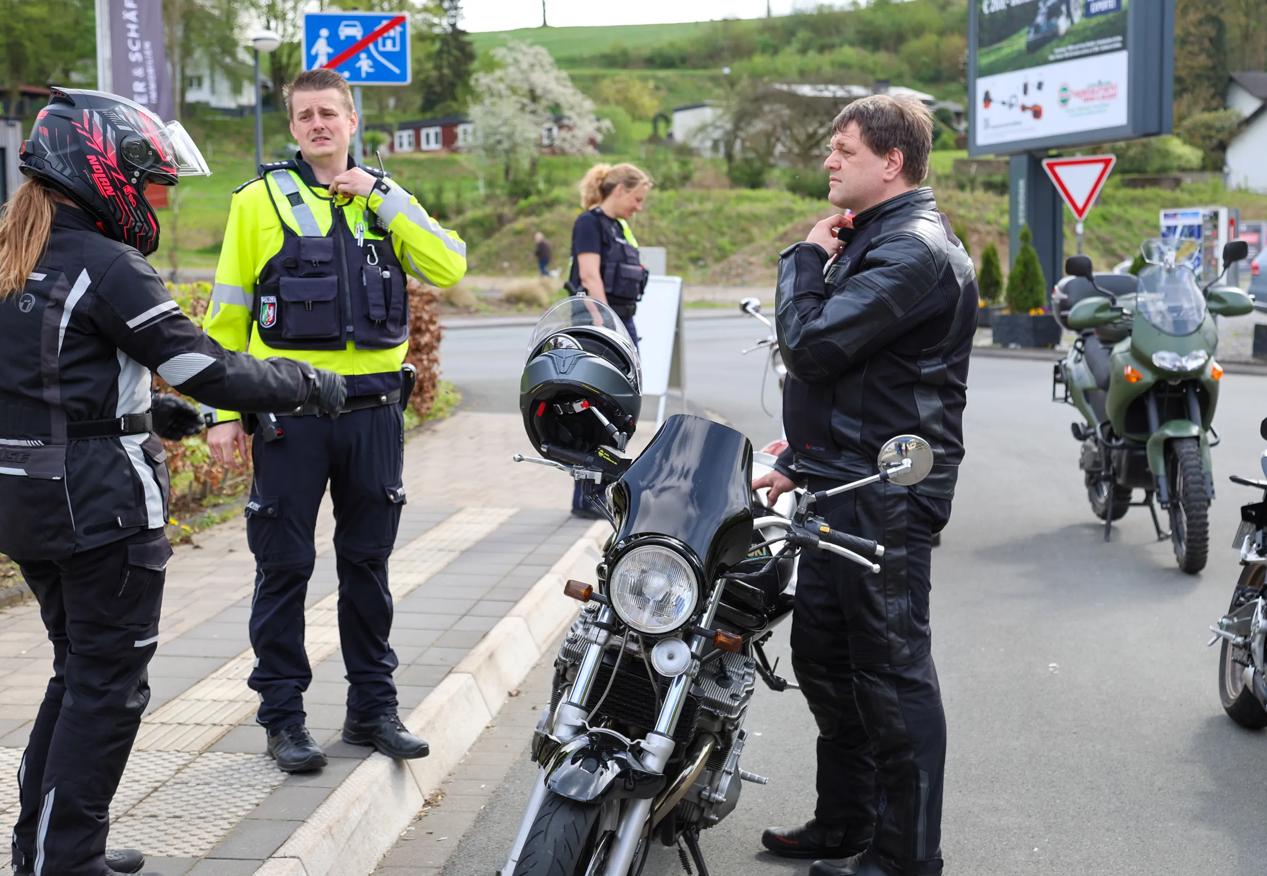 Polizisten sprechen mit einem Motorradfahrer in Schutzkleidung am Straßenrand. Im Hintergrund sind weitere Personen und Motorräder zu sehen.