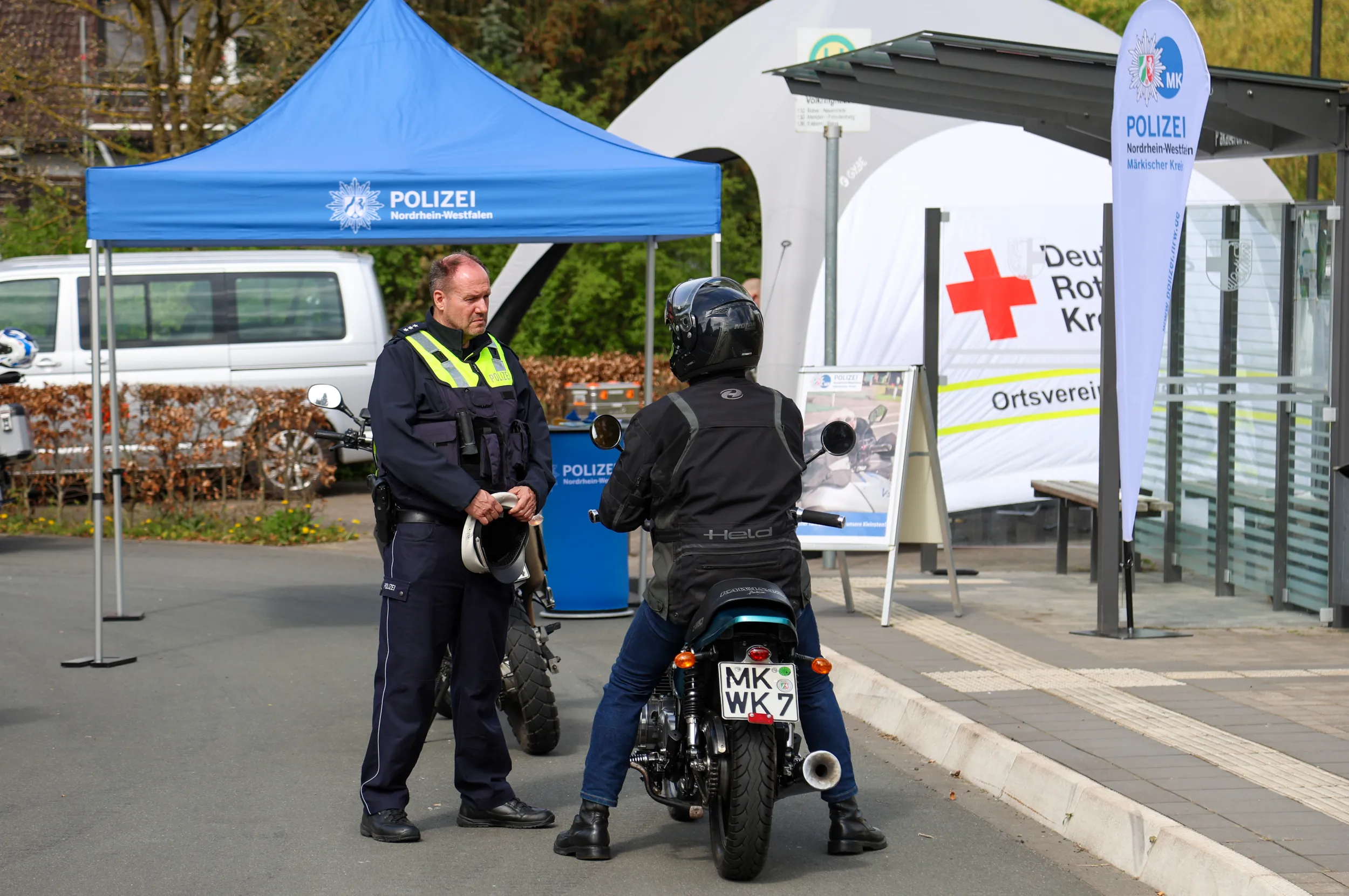 Polizist spricht Motorradfahrer während einer Kontrolle an. Im Hintergrund ein Zelt der Polizei und ein Stand des Deutschen Roten Kreuzes.