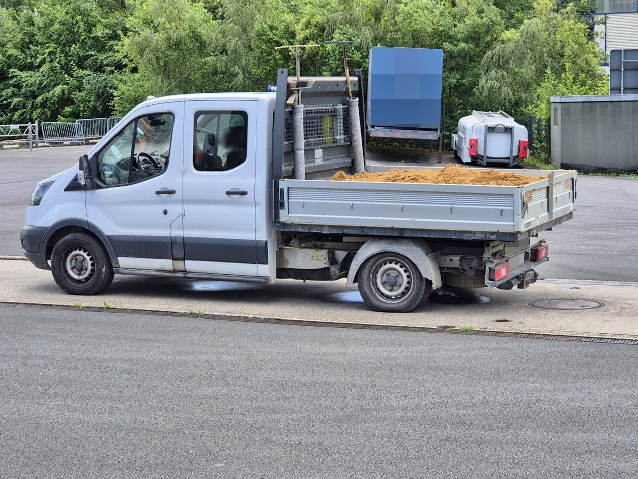 Ein weißer Klein-Lkw mit offener Ladefläche, beladen mit Sand, steht am Straßenrand. Die Ladefläche ist deutlich überladen.