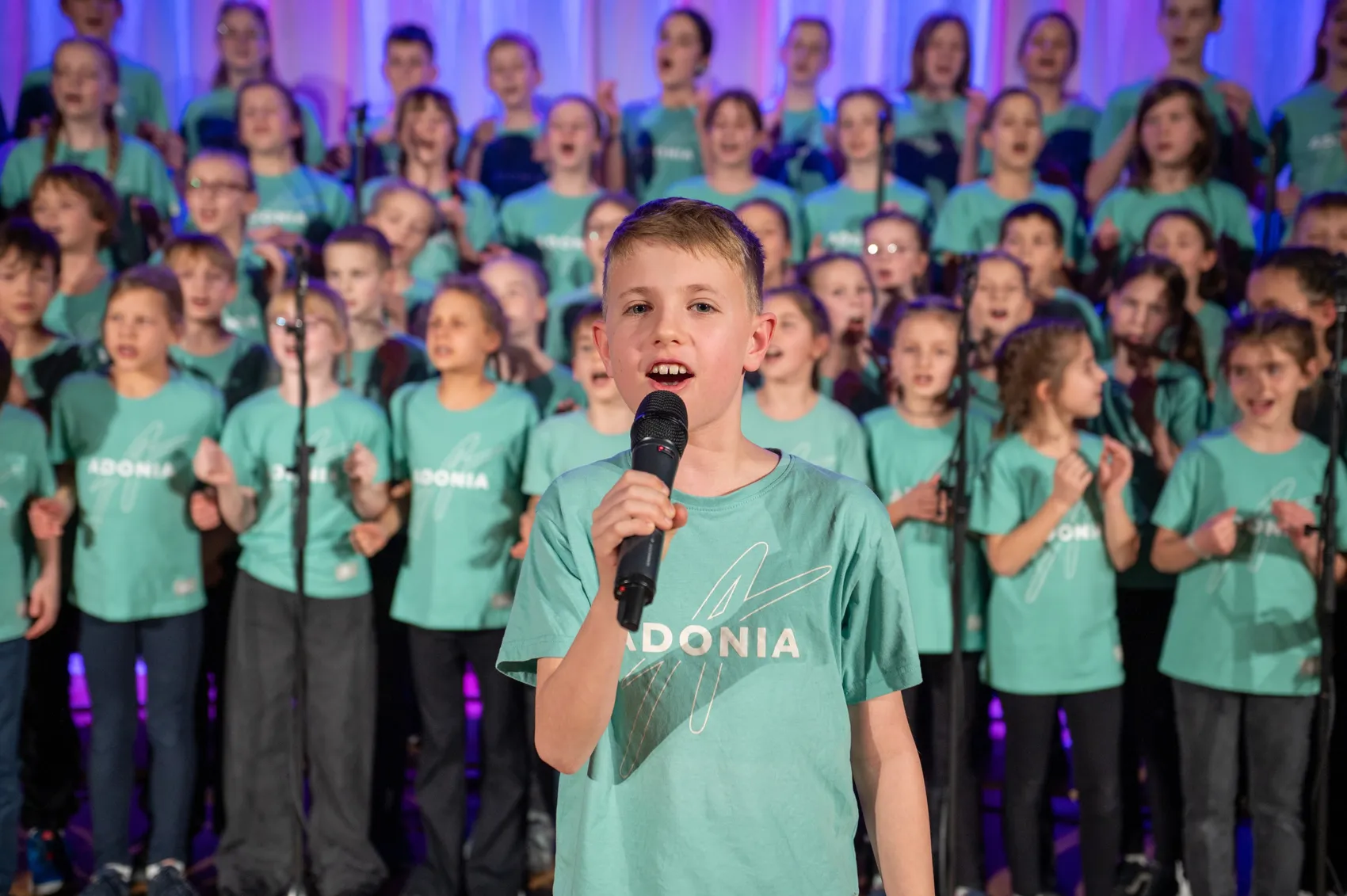 Ein Junge singt in ein Mikrofon, umgeben von einem Kinderchor in hellgrünen T-Shirts mit dem Aufdruck „Adonia“. Im Hintergrund eine Bühne mit lila Beleuchtung.