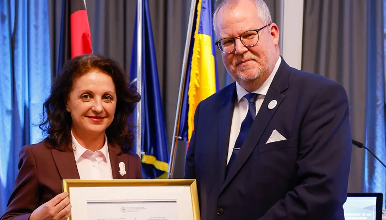 Professor Simona Mureșan überreicht Dr. Michael Klein eine Urkunde, während sie vor Flaggen und einem Bildschirm stehen.