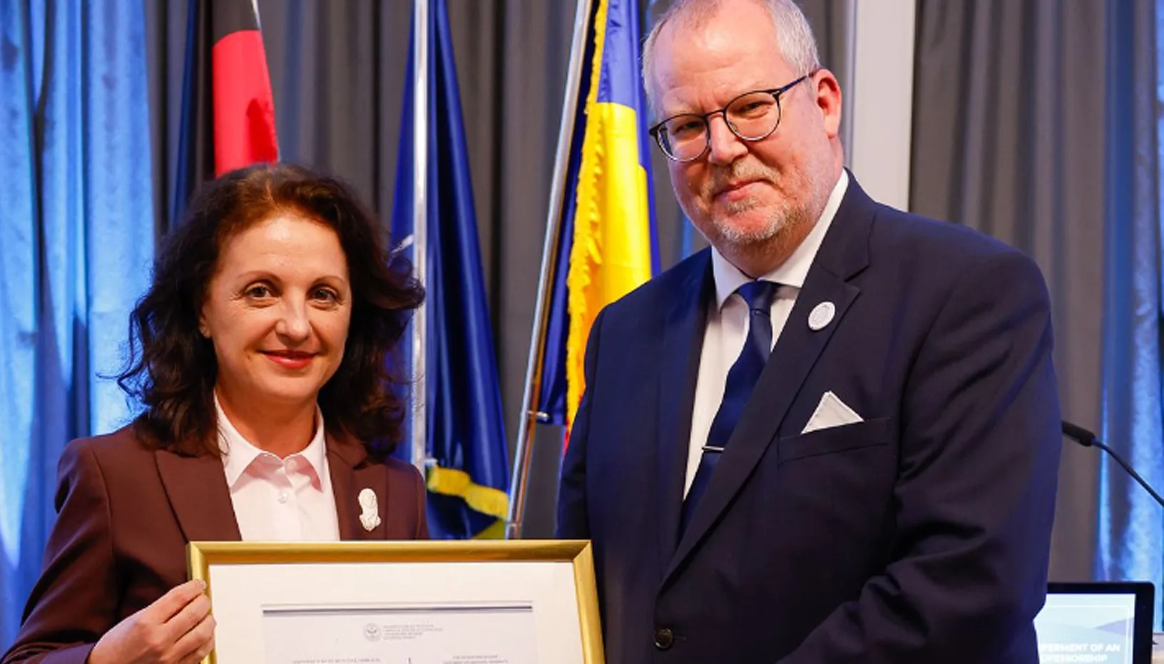 Professor Simona Mureșan überreicht Dr. Michael Klein eine Urkunde, während sie vor Flaggen und einem Bildschirm stehen.