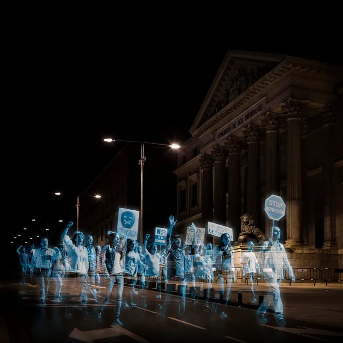 Leuchtende, transparente Hologramm-Figuren protestieren nachts mit Schildern vor dem spanischen Parlament „Congreso de los Diputados“.