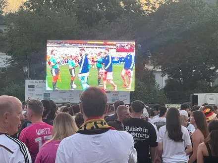 Menschen schauen Fußball auf einer Großbildleinwand im Freien. Spieler in blauen und roten Trikots sind auf dem Bildschirm zu sehen.
